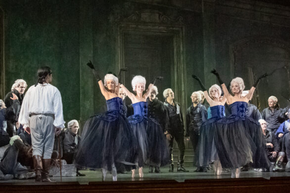 Degli attori sono in scena durante l'opera "Rigoletto". Le donne si trovano al centro del palco, in abito blu, a fianco a loro gli uomini le ammirano