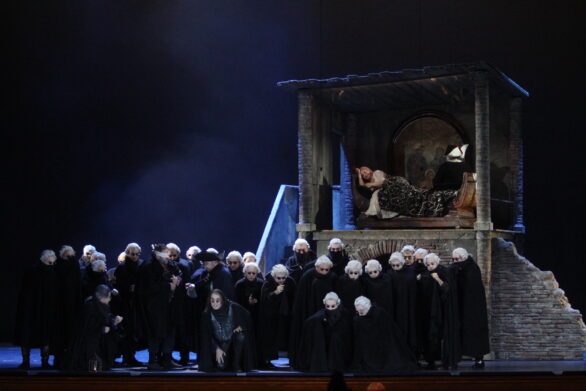 Degli attori si trovano sul palco durante l'opera "Rigoletto". Sullo sfondo si trova una scenografia di un vecchio edificio; in primo piano si trovano degli uomini vestiti di nero e parrucche bianche