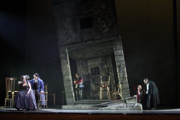 Degli attori si trovano sul palco durante l'opera "Rigoletto". Sullo sfondo si trova una scenografia di un vecchio edificio. Sulla destra e sulla sinistra si trovano dei personaggi