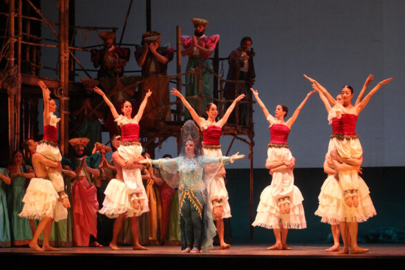 Degli attori sono sul palco immersi nei loro personaggi durante l'opera "Les pêcheurs de perles". Al centro del palco si trova una donna in abito celeste, dietro di lei altri personaggi vestiti in bianco e rosso