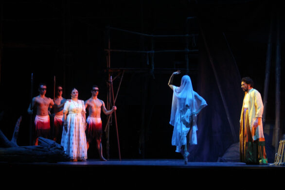 Degli attori sono in scena sul palco immersi nei loro personaggi. Sul palco si trova una scenografia molto buia
