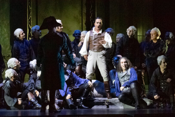 Degli attori si trovano sul palco durante l'opera "Rigoletto". Degli uomini vestiti in blu sono seduti nel palco ; un uomo in abito bianco e gilet marrone è in piedi e sta guardando un altro personaggio vestito di nero