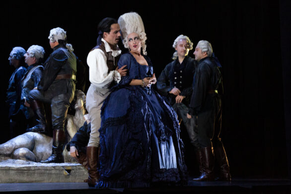 Degli attori si trovano sul palco durante l'opera "Rigoletto". Un uomo in abito bianco e grigio tiene la spalla di una donna con un abito blu e una parrucca bianca