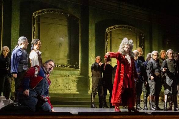 Degli attori si trovano sul palco durante l'opera "Rigoletto". Sullo sfondo si trova una scenografia di un vecchio edificio ; gli attori stanno cantando