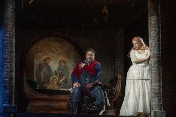 Due attori si trovano sul palco immersi nei loro personaggi durante l'opera "Rigoletto". La donna indossa un abito bianco, l'uomo invece indossa un abito rosso e blu