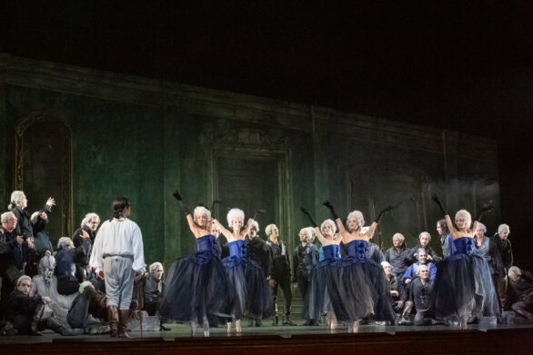 Degli attori sono in scena durante l'opera "Rigoletto". Le donne si trovano al centro del palco, in abito blu, a fianco a loro gli uomini le ammirano