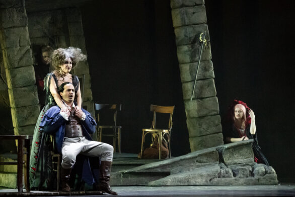 messa in scena dello spettacolo "Rigoletto". Al centro due personaggi parlano mentre una donna li spia dai lati della scenografia
