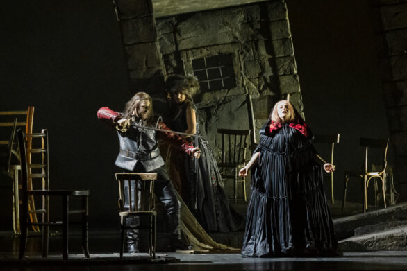 attori in scena nell'opera "Rigoletto". Una donna ha un mantello nero con cappuccio e tiene le braccia semi aperte. Un uomo la punta con una spada e una donna sullo sfondo assiste alla scena