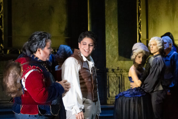 Prospettiva ingrandita del palco: degli attori sono in scena durante l'opera "Rigoletto". Un uomo vestito in bianco, con un gilet marrone sta guardando un altro personaggio