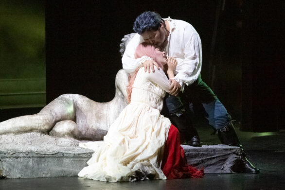 Due attori si trovano sul palco durante l'opera "Rigoletto". Una donna vestita di bianco si trova seduta sul palco e viene sorretta da un uomo