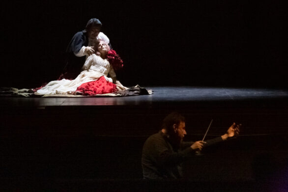 Due attori si trovano sul palco durante l'opera "Rigoletto". La donna è vestita in bianco ed è sdraiata sul pavimento, sorretta da un uomo che le accarezza i capelli