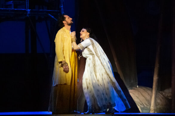 Due attori in maschera sono sul palco immersi nei loro personaggi durante l'opera "Les pêcheurs de perles". Una donna in abito bianco sta mettendo la sua mano nel petto dell'altro personaggio