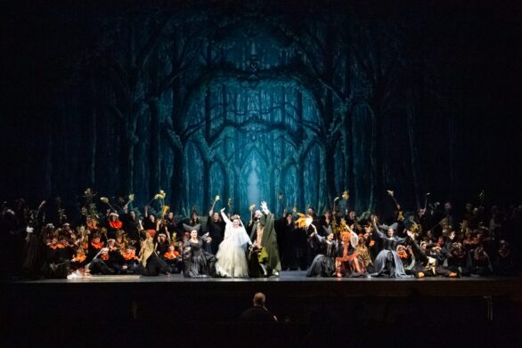 numerosi attori sul palco ne "Le allegre comari di Falstaff". Al centro della scenografia, composta da alberi, si trovano due degli attori. La donna ha un abito bianco mentre l'uomo ha un mantello bianco.