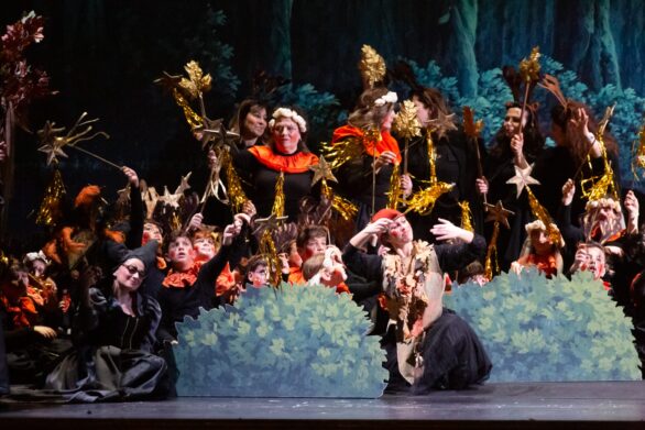 attori, adulti e bambini, in scena ne "Le allegre comari di Falstaff". La scenografia è composta da alberi e cespugli e in mano gli attori tengono delle bacchette magiche