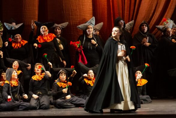 Attori in scena sul palco durante l'opera "Le allegre comari di Falstaff". Una donna in abito bianco e mantello nero si trova al centro del palco, dietro di lei ci sono dei bambini in abito nero che tengono dei fiori