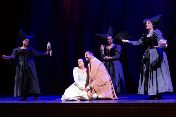 Attori in scena sul palco durante l'opera "Le allegre comari di Falstaff". Due ragazzi sorridenti si trovano al centro del palco, circondati da altri personaggi vestiti di nero