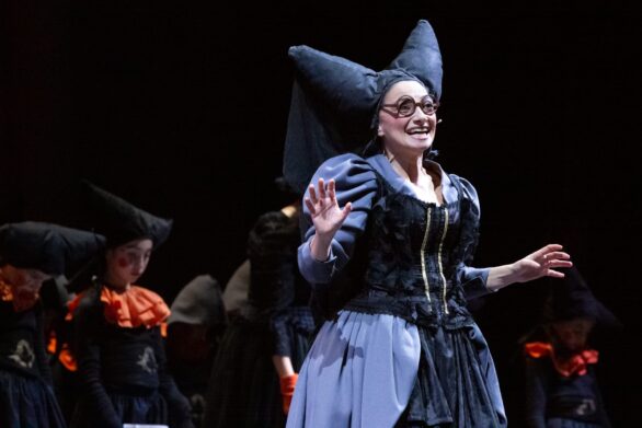 Attrice in scena sul palco durante l'opera "Le allegre comari di Falstaff". Indossa un abito nero e un cappello stravagante
