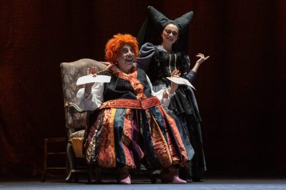 Attori in scena sul palco durante l'opera "Le allegre comari di Falstaff". Un uomo in maschera è seduto su una poltrona, al suo fianco si trova una donna in abito nero.