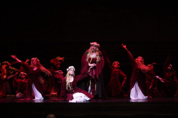 delle figure femminili vestite di rosso, con una corona di fiori in testa ballano e cantano sul palco