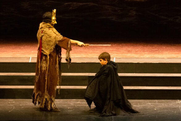 Scena in atto sul palco. Un uomo con una maschera dorata sta toccando con una spada la testa di un bambino.