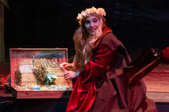 primo piano di un'attrice sul palco. E' seduta su uno scalino della scenografia e sta prendendo in mano una boccetta di vetro che si trovava all'interno di una cassetta di legno