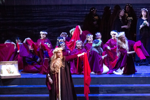 scena ingrandita del palco. Una donna con un mantello lungo si trova al centro del, dietro di lei si trovano dei bambini immersi nei loro personaggi