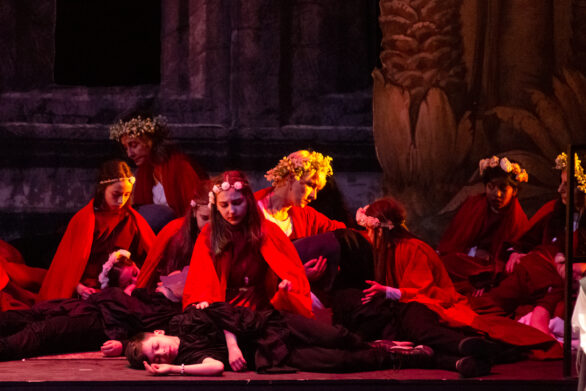 diverse donne e bambine della compagnia teatrale durante la scena di uno spettacolo. Alcune di loro indossano un mantello rosso e una corona di fiori e si prendono cura d altri distesi per terra