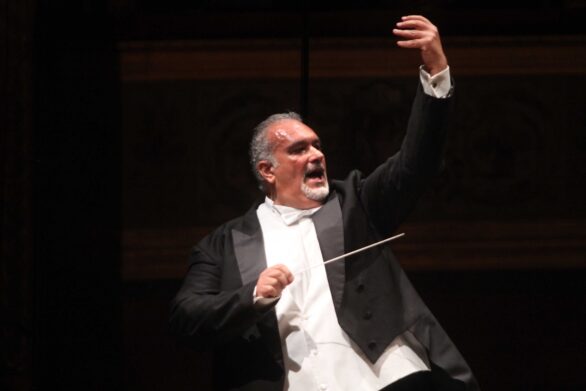 Il direttore d’orchestra, ripreso frontalmente, mentre guida con gesti energici e precisi l’orchestra.