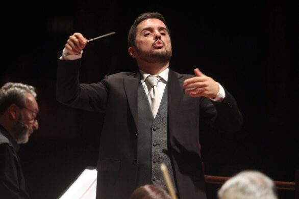 Il direttore d’orchestra Alberto Maniaci, in abito scuro, è ripreso frontalmente mentre dirige con la bacchetta in mano. Sullo sfondo l’attore Fabrizio Lupo sta recitando.