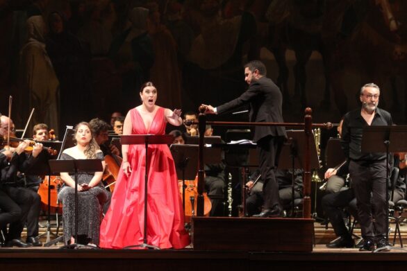 Soprano Maria Cristina Napoli, vestita di rosso, con braccio sinistro sollevato, concentrata sull'esecuzione. Alla sua destra il direttore d’orchestra.