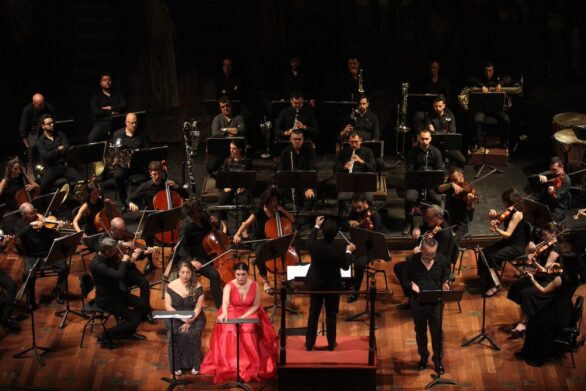 Veduta del palco dall’alto, con l’orchestra in esecuzione. Il direttore, di spalle, guida l’orchestra. In primo piano due artiste vestite di nero e rosso.