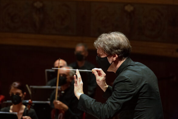 Profilo del direttore d’orchestra Marc Albrecht con bacchetta in mano. Sullo sfondo i violinisti