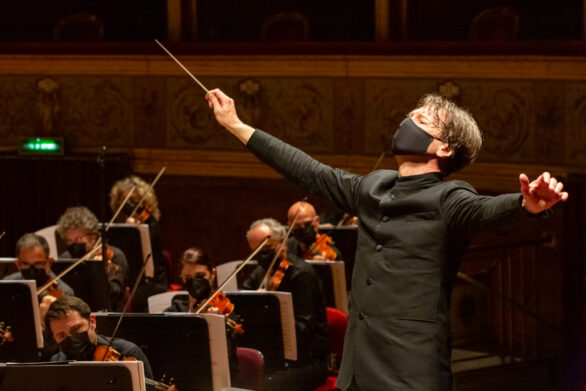 Primo piano del direttore d’orchestra Marc Albrecht a braccia aperte, sullo sfondo i violinisti
