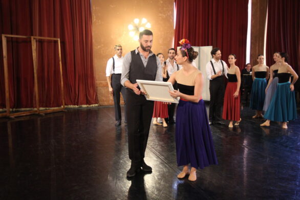 Una ballerina e un uomo tengono insieme una cornice in sala. Nello sfondo altri ballerini