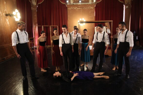 Un gruppo di danzatori in abiti formali, sono disposti in cerchio attorno a una figura femminile distesa sul pavimento, suggerendo una scena drammatica all’interno di un ambiente elegante.