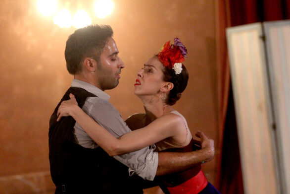 la coppia balla, con espressioni intense e ravvicinate. La donna , Frida, indossa un accessorio floreale nei capelli e guarda con intensità il suo partner, mentre l’uomo è concentrato, evidenziando un’atmosfera di passione e coinvolgimento emotivo.