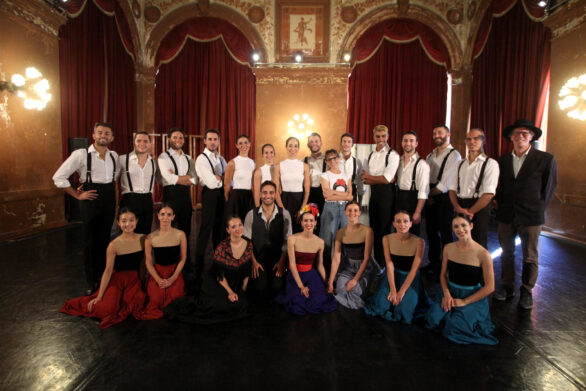Gruppo di ballerini e ballerine in posa sul palco. Gli uomini indossano camicie bianche e bretelle nere, le donne abiti lunghi colorati.